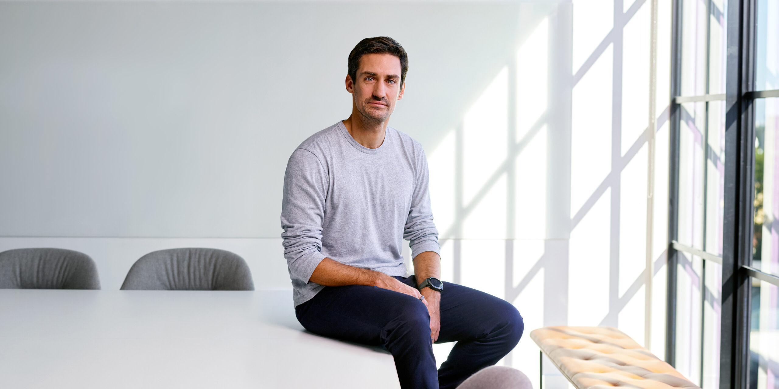 Executive in a bright modern office, leaning against a desk with floor-to-ceiling windows and city views behind him.